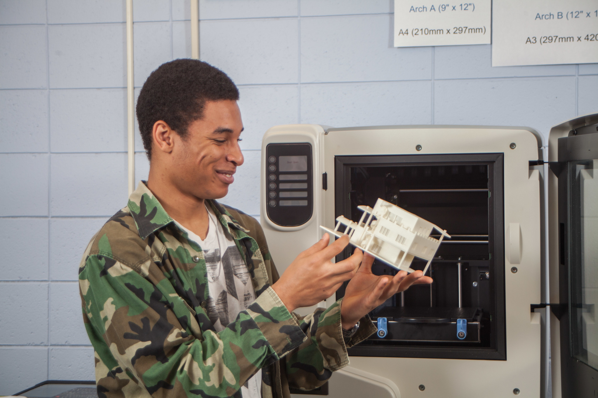 A male student is taking a white ABS plastic part out of a 3D printer. The part appears to be a house.
