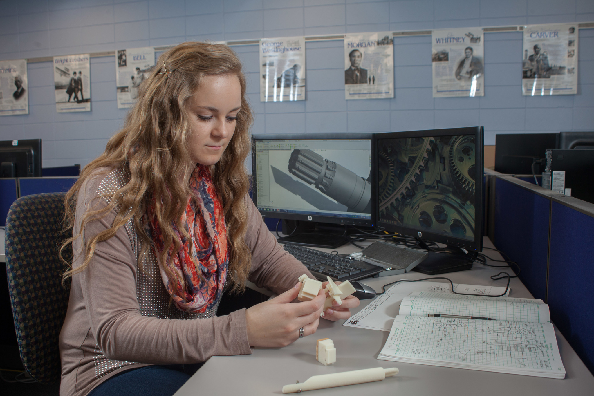 A female student is sitting at a workstation designing a part with technical images on the monitor screens behind her.
